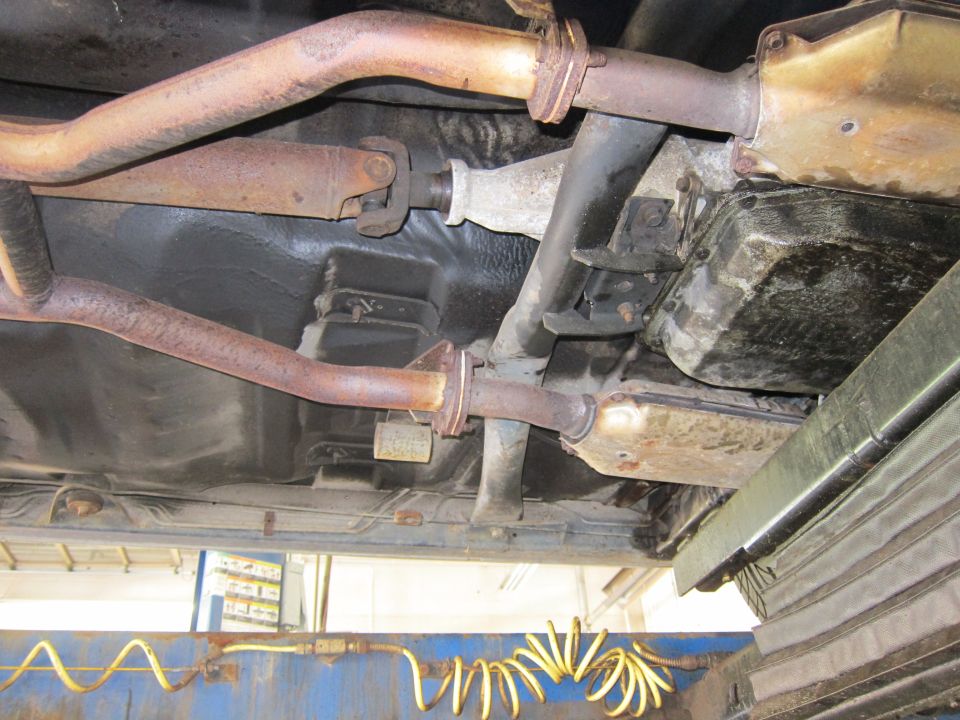 Underside view of a vehicle's exhaust system and drivetrain components, showing rust on the pipes and a greasy transmission pan