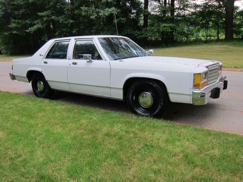 1991 Ford LTD Crown Victoria - White sedan, likely from the early 1980s, parked on a grassy area beside a paved road.