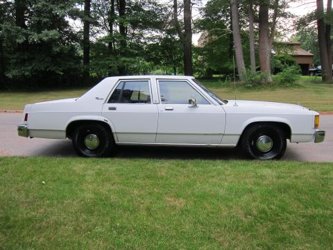 1991 Ford LTD Crown Victoria - A white sedan is parked on a grassy area next to a road, with trees and a house in the background.