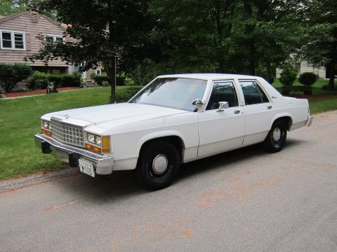 1991 Ford LTD Crown Victoria - A white 1980s Ford sedan is parked on a street in front of a house and trees.