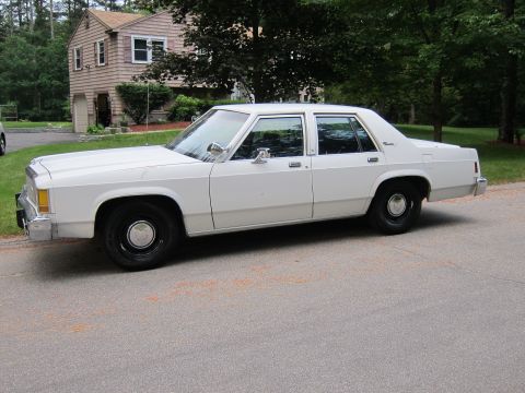 1991 Ford LTD Crown Victoria - This white sedan, likely from the 1980s, is seen from the side with a house and trees in the background.