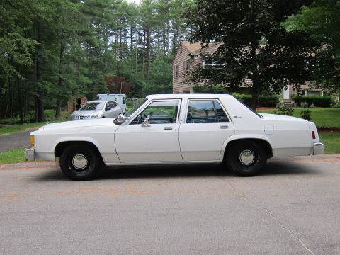 1991 Ford LTD Crown Victoria - Full driver-side profile of a white Ford LTD Crown Victoria four-door sedan on an asphalt driveway, with pine woods and a shingled house be…