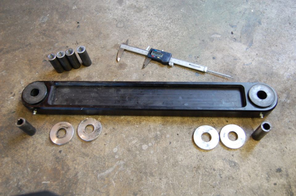 Black suspension control arm with four metal spacers, two washers, two bolts, and digital calipers on a concrete surface