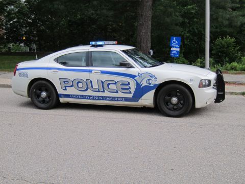 1991 Ford LTD Crown Victoria - Side profile of a white-and-blue University of New Hampshire police Dodge Charger marked POLICE 911 parked on a street.