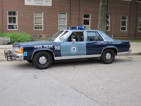 1991 Ford LTD Crown Victoria - Side profile of a two-tone blue Massachusetts State Police sedan marked 90E parked in front of a brick building.