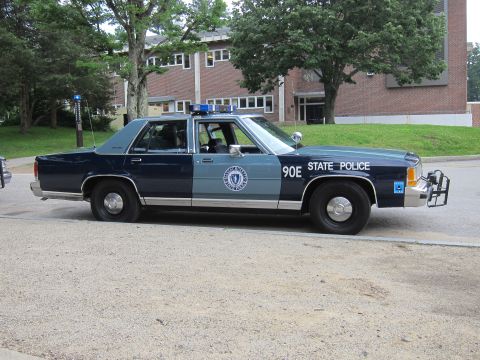 1991 Ford LTD Crown Victoria - Side profile of a navy-and-gray Massachusetts State Police sedan lettered '90E STATE POLICE' with a push bar, parked on a gravel lot.