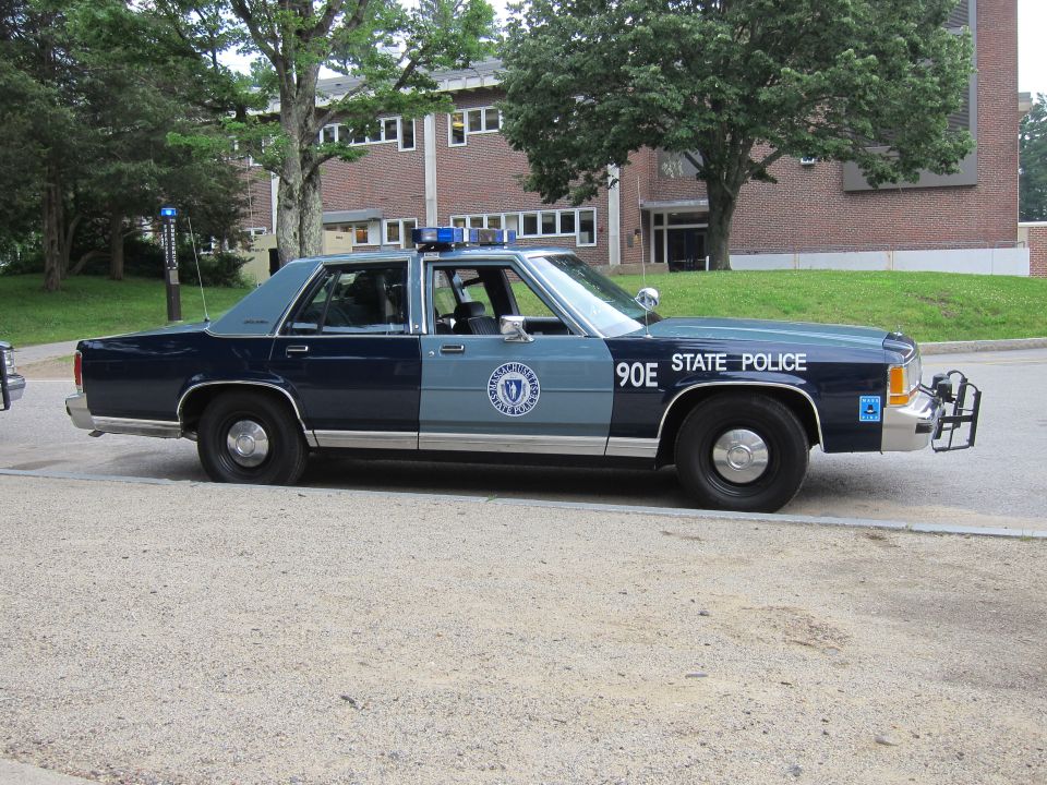Navy and gray Massachusetts State Police Ford sedan side profile on gravel lot