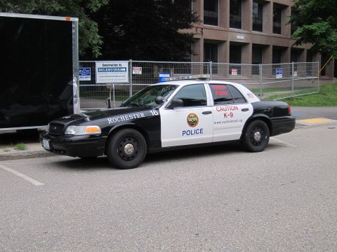 1991 Ford LTD Crown Victoria - Front quarter view of a black-and-white Rochester Police Ford Crown Victoria K-9 cruiser parked by a chain-link fence and brick building.