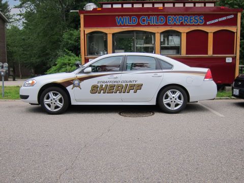 1991 Ford LTD Crown Victoria - white Chevrolet Impala marked 'STRAFFORD COUNTY SHERIFF' with gold-and-brown striping, parked before a red 'Wild Child Express' kiosk.