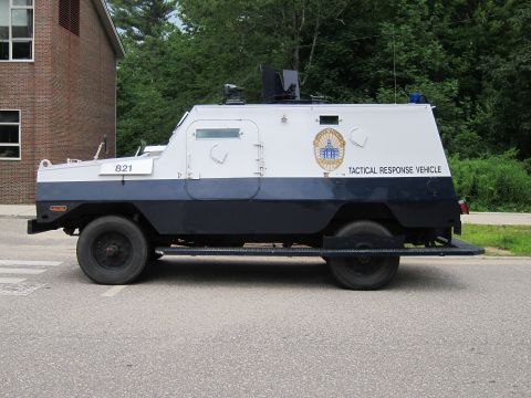 1991 Ford LTD Crown Victoria - Full side view of a white-and-navy armored police 'TACTICAL RESPONSE VEHICLE' numbered 821 with a turret hatch, parked by a brick wall.