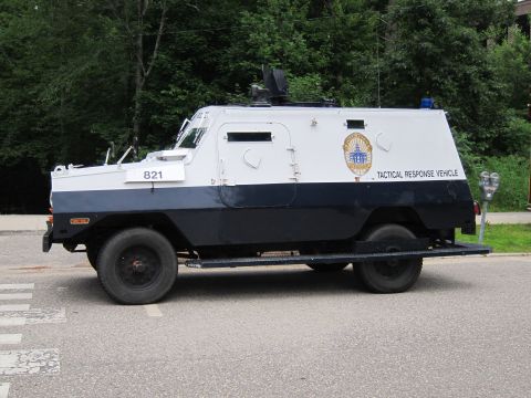1991 Ford LTD Crown Victoria - Side view of a white-and-navy armored police 'TACTICAL RESPONSE VEHICLE' numbered 821 with roof turret, parked on a road by trees.