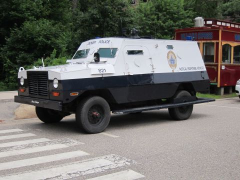 1991 Ford LTD Crown Victoria - white-and-navy Dover Police armored 'TACTICAL RESPONSE VEHICLE' numbered 821 with grille and turret, parked near a crosswalk and red kiosk.