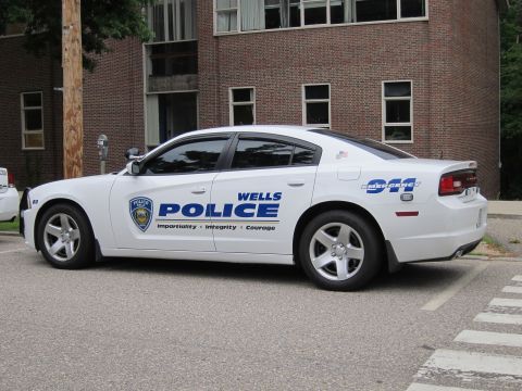 1991 Ford LTD Crown Victoria - white Dodge Charger marked 'Wells Police' with a blue shield decal parked alongside a brick building.