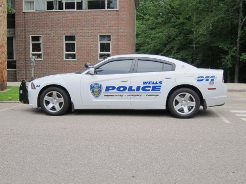 1991 Ford LTD Crown Victoria - Passenger-side view of a white Wells Police Dodge Charger with push bar, parked near a brick building.