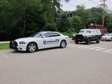 1991 Ford LTD Crown Victoria - white Wells Police Dodge Charger towing or leading a dark armored truck along a crosswalk on a tree-lined road.