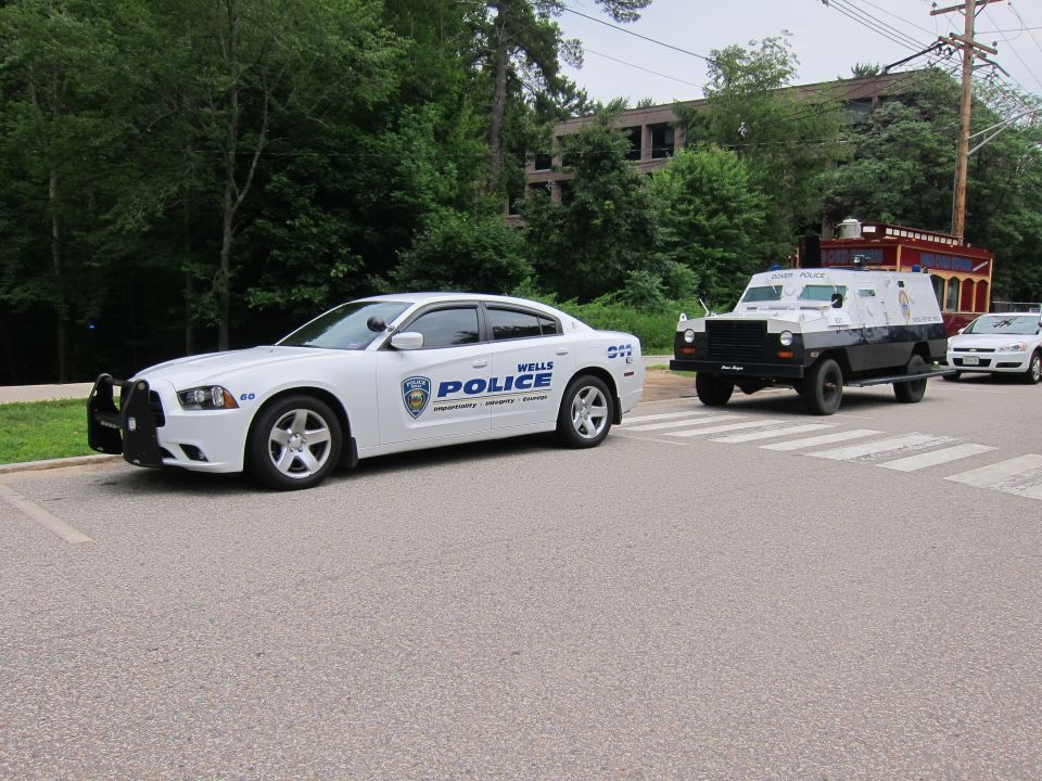 White Wells Police Charger towing a dark armored truck on a wooded road