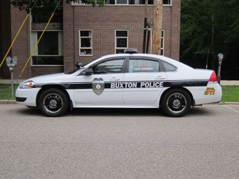 1991 Ford LTD Crown Victoria - Side view of a silver and black Chevrolet Impala marked 'Buxton Police' with 'Dial 911' decal, parked on a street.