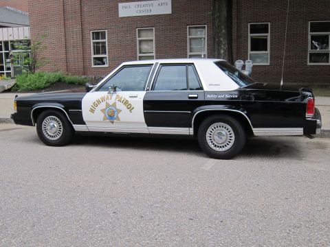 1991 Ford LTD Crown Victoria - Side view of a black and white Ford LTD Crown Victoria marked 'Highway Patrol' with star emblem, parked by a building.