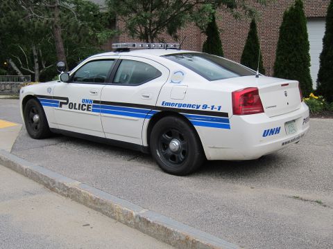 1991 Ford LTD Crown Victoria - white Dodge Charger marked UNH Police with blue striping and Emergency 9-1-1 lettering, parked at a curb by a brick building.