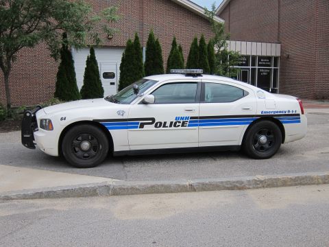 1991 Ford LTD Crown Victoria - Driver-side profile of a white Dodge Charger UNH Police cruiser with blue stripe and push bar, parked before a brick building.