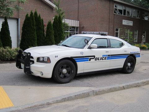 1991 Ford LTD Crown Victoria - white Dodge Charger UNH Police cruiser, unit 64, with brush guard and roof lightbar parked along a curb at a brick building.