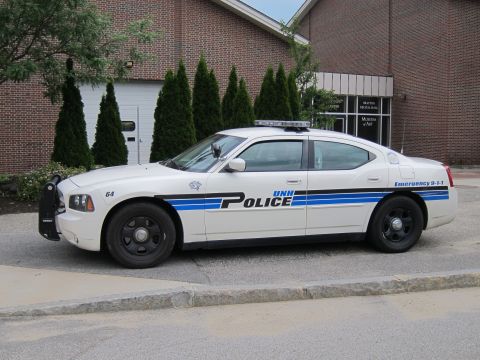 1991 Ford LTD Crown Victoria - white Dodge Charger UNH Police cruiser, unit 64, parked along a curb in front of a brick building with evergreens.