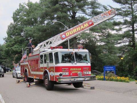1991 Ford LTD Crown Victoria - red Durham Ladder fire truck with an extended aerial ladder, parked by a road with a firefighter atop and a flag.