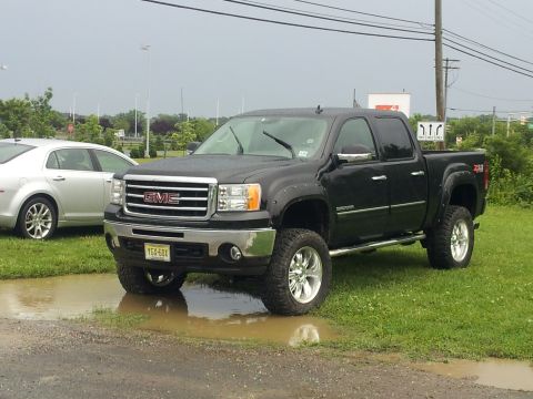 1978 Chevrolet C/K Pickup - A black GMC Sierra pickup truck is parked on grass next to a silver sedan, with a puddle in front of it.