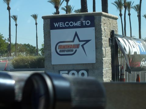 2003 Ford Crown Victoria - 'Welcome to Irwindale Event Center' sign with a star logo and address numbers, framed by palm trees and a dashboard gauge in the foreground.
