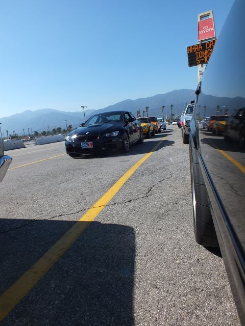 2003 Ford Crown Victoria - black BMW coupe waits in a staging lane lined with cars at a drag strip, with mountains and a Toyota sign in the background.