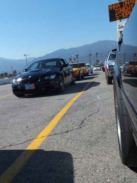 2003 Ford Crown Victoria - black BMW coupe at the front of a line of cars at a drag strip, beneath an 'NHRA Drags Tonight' sign with mountains behind.
