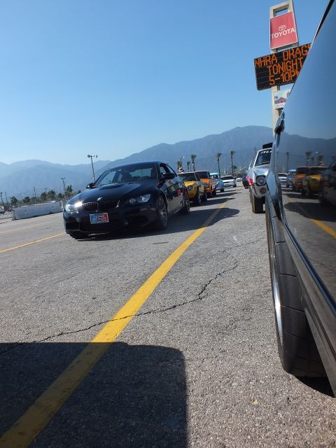 2003 Ford Crown Victoria - black BMW coupe waits in a line of cars at a drag strip, with a Toyota sign and mountains in the background.