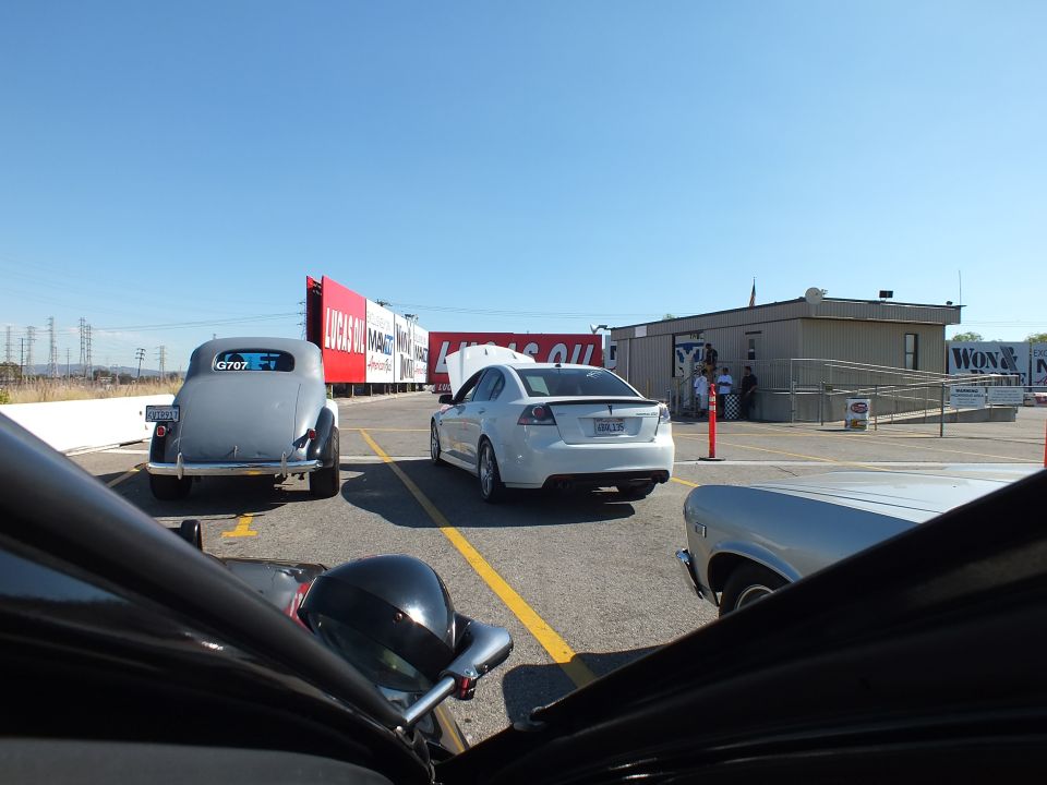 Vintage hot rod and silver sedan parked at a drag strip near a tower building