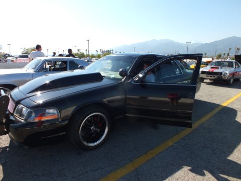 2003 Ford Crown Victoria - black full-size sedan with a push-bar grille guard and open door, parked among classic cars at a sunny lot with mountains behind.