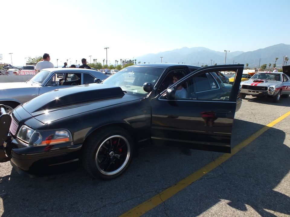 Black sedan with grille guard and open door at a car-show lot, mountains behind