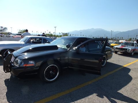 2003 Ford Crown Victoria - black full-size sedan with a chrome push-bar and open driver's door, parked among muscle cars in a paved lot with mountains behind.