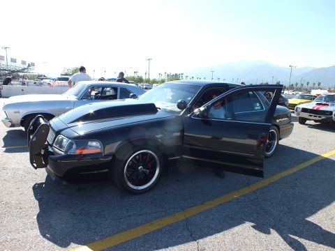 2003 Ford Crown Victoria - black full-size sedan with push-bar grille guard and open door, parked beside classic cars in a sunny lot with mountains in the distance.