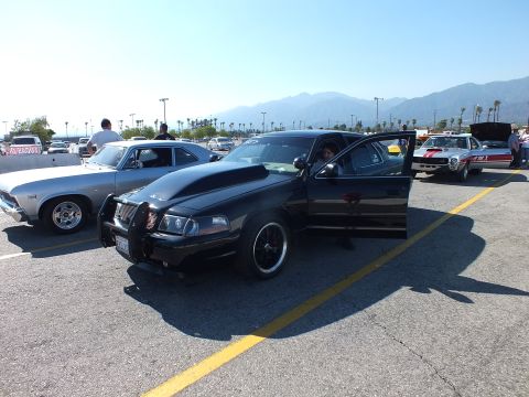 2003 Ford Crown Victoria - black full-size sedan with a chrome grille guard parked beside a silver classic muscle car in a sunny lot with mountains behind.