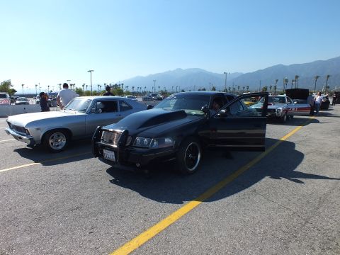 2003 Ford Crown Victoria - black full-size sedan with a push-bar grille guard parked alongside a silver classic car in a paved lot with mountains in the background.