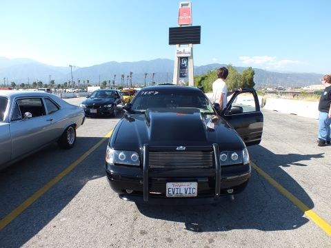 2003 Ford Crown Victoria - black full-size sedan with a chrome push-bar and 'EVIL VIC' plate viewed head-on, with people and a Toyota sign in the background.