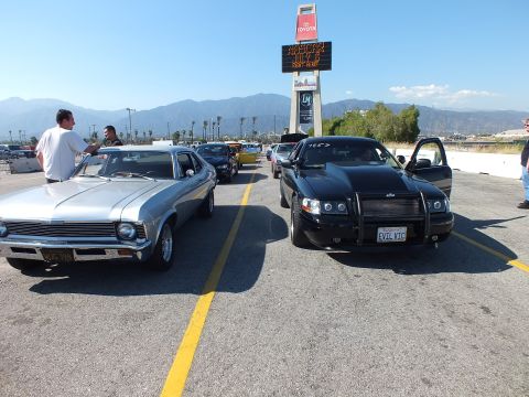 2003 Ford Crown Victoria - silver classic muscle car and a black sedan with grille guard and 'EVIL VIC' plate parked facing each other, people standing nearby.