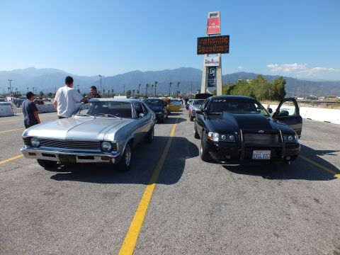 2003 Ford Crown Victoria - silver classic muscle car beside a black sedan with grille guard and 'EVIL VIC' plate, with people and a Toyota sign behind.