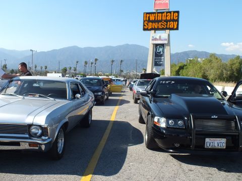 2003 Ford Crown Victoria - silver classic muscle car and a black 'EVIL VIC' sedan in a line of cars beneath an Irwindale Speedway sign with mountains behind.