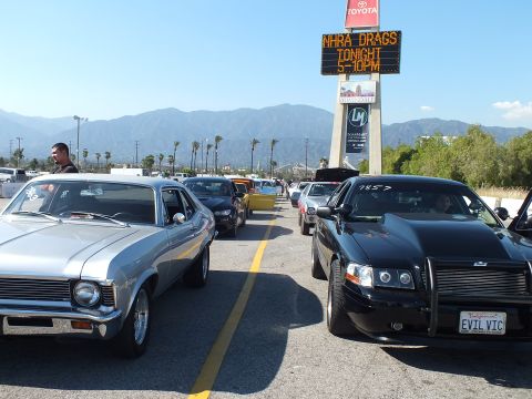2003 Ford Crown Victoria - silver classic muscle car and a black 'EVIL VIC' sedan in a line of cars under an 'NHRA Drags Tonight' sign with mountains behind.