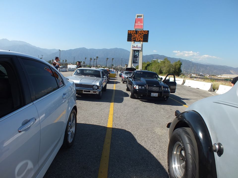 View down a line of cars at a drag strip with white sedan and hot rod in foreground