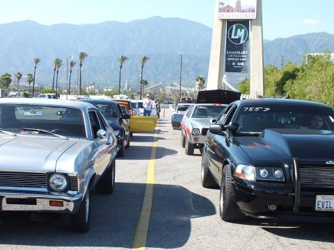 2003 Ford Crown Victoria - silver Chevrolet Nova and a black sedan with a personalized 'EVIL' plate sit in the staging lanes beside an Irwindale LoanMart tower sign.