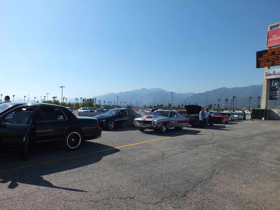Wide staging-area view of a black sedan, striped muscle car, and open-hood cars under a tower sign