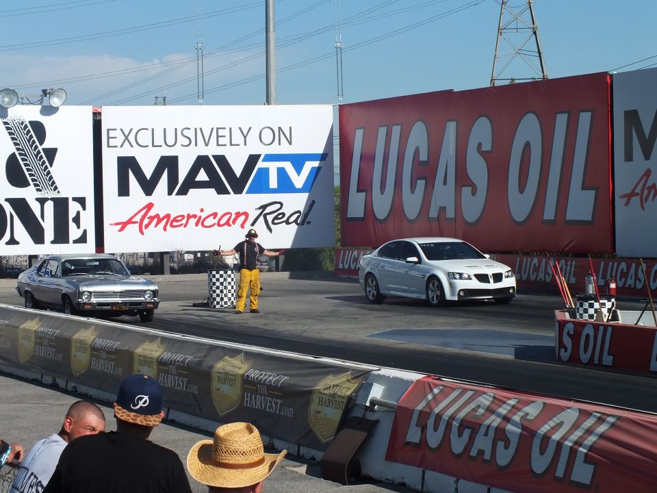 Two cars at the drag-strip start line with a starter, before MavTV and Lucas Oil wall banners