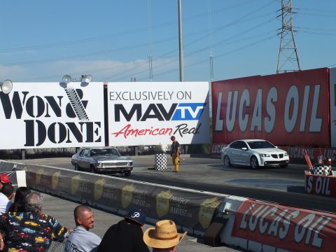 2003 Ford Crown Victoria - silver muscle car and a white Pontiac G8 sit at the start line under 'Won & Done' and MavTV banners while spectators watch from the stands.