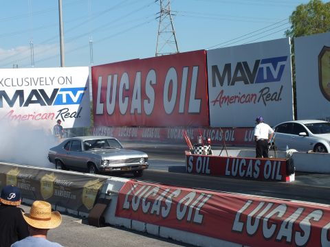 2003 Ford Crown Victoria - silver muscle car lays down a smoky burnout at the start line as a white sedan waits, with MavTV and Lucas Oil banners on the wall.